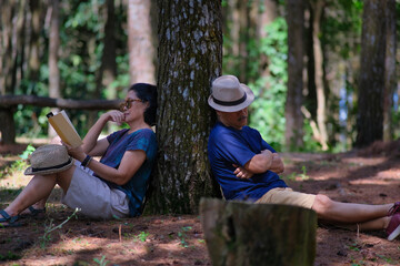 A woman leaning against a tree trunk reading a book; a man leaned on the other side of the tree while sleeping - obrazy, fototapety, plakaty