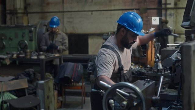 Medium shot of Middle Eastern male industrial engineer in hard hat using special hardware tools while repairing mechanism in lathe machine during night shift at metalworks factory