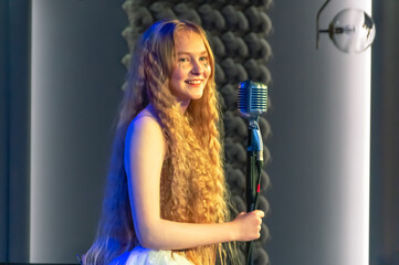 A young girl with blonde curly hair sings into a retro microphone. Performance of a girl with long hair on stage. The performance of the song at the concert. A smile on the girl's face.