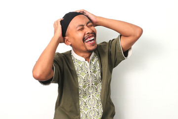 Overjoyed Indonesian Muslim man in koko shirt and peci grabs his head in a gesture of excitement,...