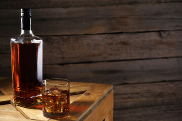 Whiskey with ice cubes in glass and bottle on wooden crate, space for text