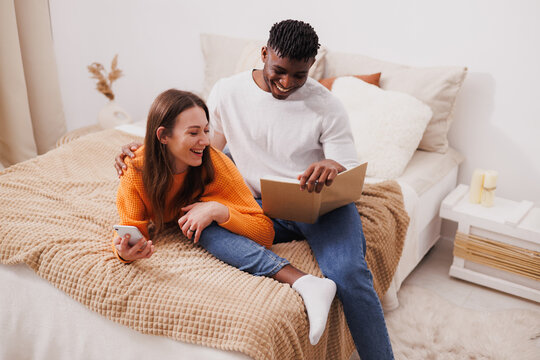 Cheerful multiethnic couple reading book and using smartphone on bed - Powered by Adobe