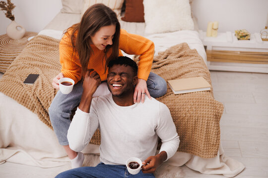 Positive multiethnic couple with coffee cups having fun in bedroom