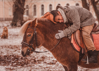 a boy sitting in a saddle lies on the neck of a pony and smiles thoughtfully