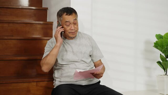 Footage handheld medium shot, Concerned Asian retired man sits on stairs, worried about home expenses. holding paper bills and smartphone in hand, Real-life financial stress concept.
