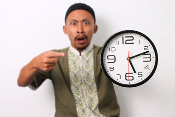 A shocked Indonesian Muslim man in koko and peci emphatically points at a clock, realizing he's late for his sahur meal during Ramadan. Isolated on a white background - obrazy, fototapety, plakaty