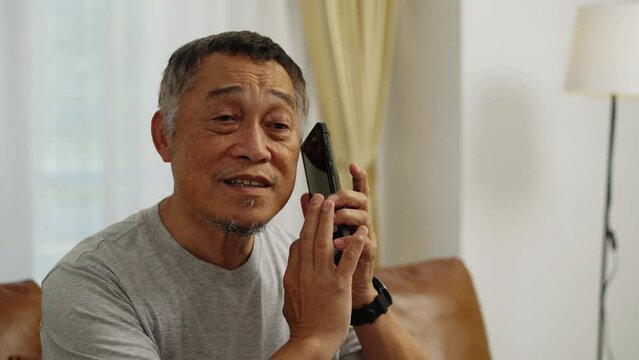 Footage handheld close up shot, Asian mature man who lost his vision was using his finger to scroll the screen of his mobile phone to listen voice, he smiled and laughed happily in the living room.