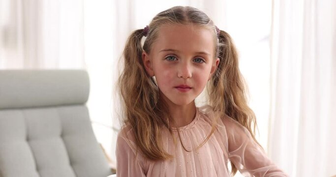 Serious little girl of five years old, looking sternly in thought at the camera with two ponytails in a pink dress, close-up portrait standing at home