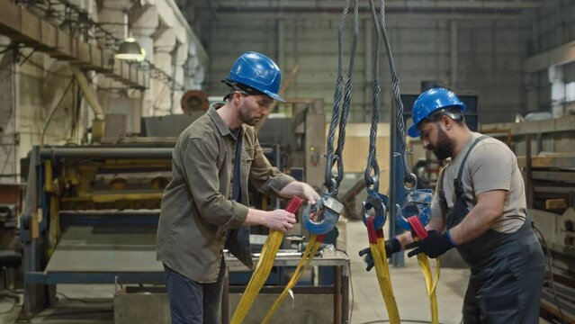 Tilt up shot of two ethnically diverse workmen in hard hats fastening industrial lifting belt to crane to lift up heavy palette from floor at industrial metalworks factoryTilt up shot of two ethnicall