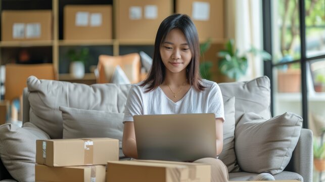 Asian business woman on sofa using a laptop computer checking customer order online shipping boxes at home.