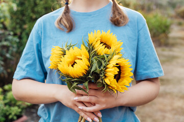 Focus on fresh sunflowers bouquet in hands of young female farmer on the green trees background. Growing flowers. Rural, Cottage core lifestyle. Selective focus.