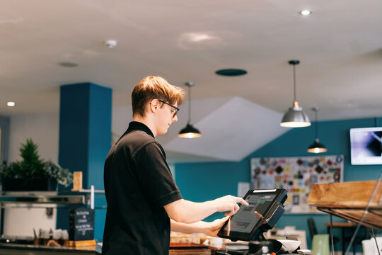 Young waiter serving customer at cash point in cafe. Man working with POS terminal. Cashier, barista checking for payment receipt. Hospitality, server and preparing a slip at the till in coffee shop