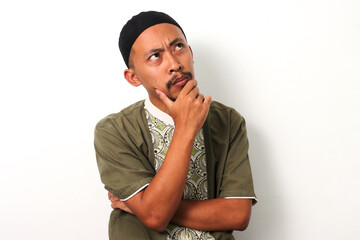 A pensive Indonesian Muslim man in koko and peci rests his hand on his chin in contemplation during Ramadan. Isolated on a white background - obrazy, fototapety, plakaty