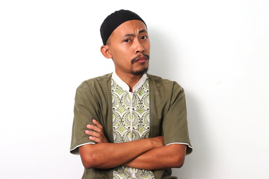 Thoughtful Indonesian Muslim man in koko shirt and peci crosses his arms, looking at the camera with a doubtful or confused expression. Isolated on White background