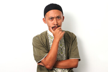 A pensive Indonesian Muslim man in koko and peci rests his hand on his chin, gazing thoughtfully at...