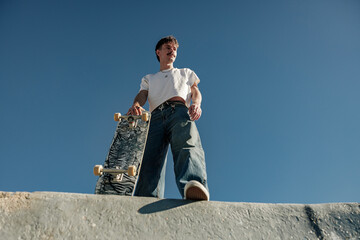 Low angle of happy male skateboarder holding skateboard standing at skate park in sunny day © Kostiantyn