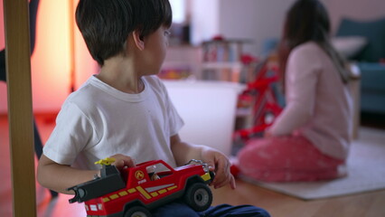 Little boy playing with fire truck vehicle toy on floor with sister in background. Focused child holding object in hand