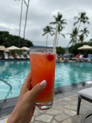 A photo taken lounging on a chair by the pool, featuring two orange cocktails on the table, with the outdoor swimming pool in the background. The sky is partly cloudy, yet warm in feeling.
