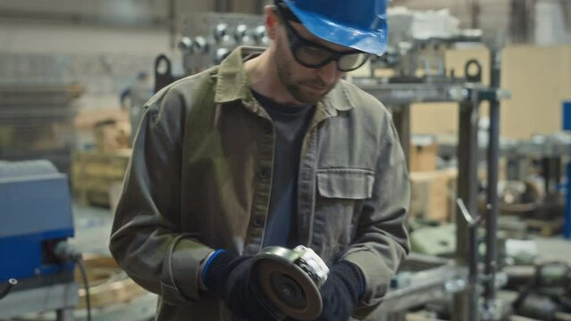 Tilt shot of male metalworker in safety glasses and hard hat using grinding tool on metal construction while working at industrial factory