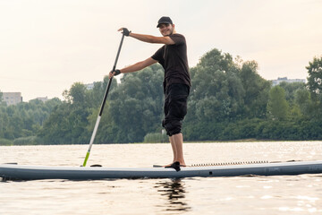 Young man has fun gliding on paddle board over urban lake. Sport enthusiast spends vacation engaged...