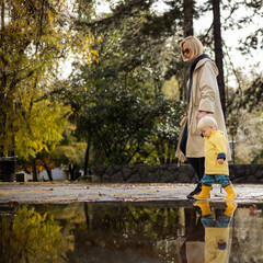 Small bond infant boy wearing yellow rubber boots and yellow waterproof raincoat walking in puddles on a overcast rainy day holding her mother's hand. Mom with small child in rain outdoors - 748791551