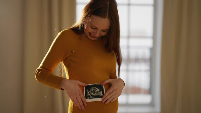 Pregnant woman looking ultrasound report. Beautiful pregnant female watching her ultrasound report and touching her abdomen, admiring sonography picture of her unborn baby. Expecting baby concept.