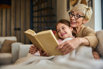 A lovely grandma reading a book story for her grandchild, sitting on the couch, wrapped in a...