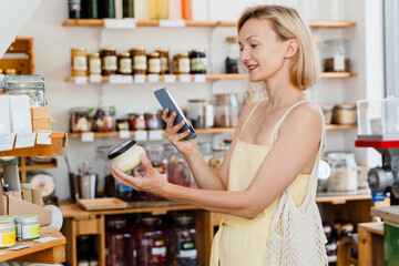 Thoughtful woman using smartphone buying products at store. Сonscious shopping and retail of organic food and eco goods at local small shop.