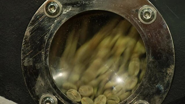 Green coffee beans fall down into roasting machine with round glass window in shop closeup. Process natural product for energy drink