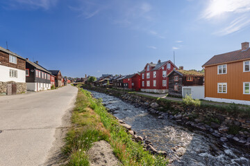 Fototapeta na wymiar A road runs parallel to the Glomma River in Roros, flanked by a charming row of traditional wooden houses basking in the Nordic sunlight