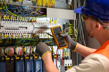 electrician in blue helmet, yellow glasses and orange vest measures electric current with digital multimeter in distribution box-blurred background