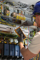 an electrician in a blue helmet, yellow glasses and an orange vest measures electric current with a digital multimeter on a distribution box
