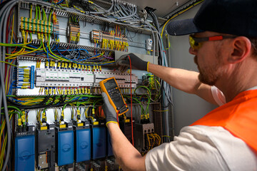 an electrician in a cap, yellow glasses and an orange vest measures electric current with a digital...