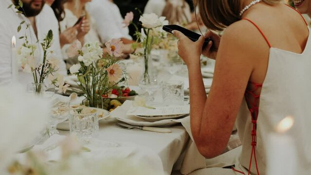 Woman taking a photo of wedding table. Close up shot of wedding table in the greenhouse served and decorated with pastel flowers for minimalistic natural wedding dinner. Wedding guest gathering. 
