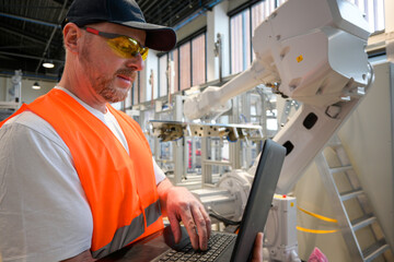 an engineer in a vest is programming a robot on an assembly line using a laptop