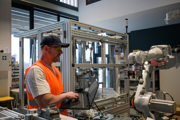 an engineer in a vest is programming a robot on an assembly line using a laptop