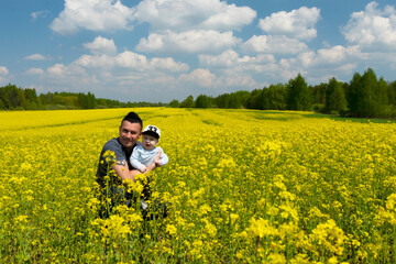Naklejka na ściany i meble Handsome dark-haired man, dad holds his little son in his arms in a rapeseed field. Copy space.