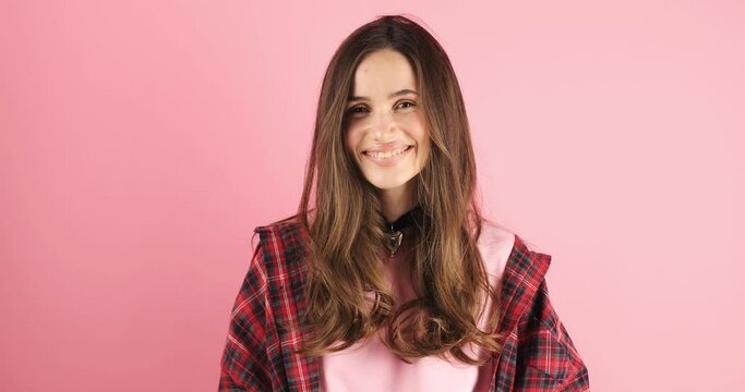 Playful happy contented brunette woman wear red plaid shirt blinking eye, looking at camera with toothy smile, winking and flirting, expressing optimism. Indoor studio shot isolated on pink background