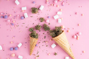 Dry buds of medical marijuana lie on waffle ice cream cones on a pink background.  There are candies and marshmellos around.  Alternative medical cannabis treatment