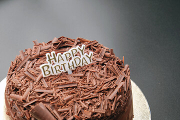 A dark black forest chocolate cake with a Happy Birthday sign sits on a black background