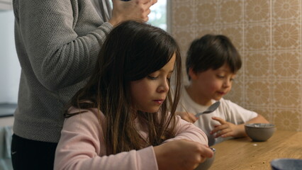 Pensive little girl eating yogurt from bowl in kitchen with mother and little brother in the background. Domestic authentic lifestyle scene of family eating breakfast early in the morning