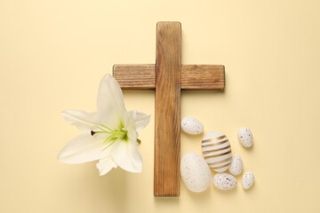 Wooden cross, painted Easter eggs and lily flower on pale yellow background, flat lay