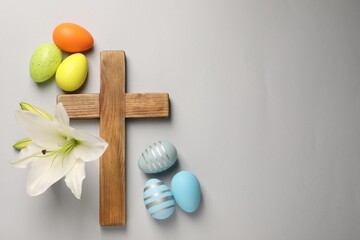 Wooden cross, painted Easter eggs and lily flowers on grey background, flat lay. Space for text