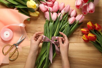 Woman making beautiful bouquet of fresh tulips and ribbon at wooden table, top view
