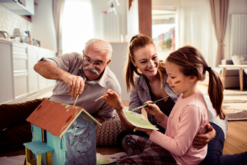 Grandfather, mother and granddaughter painting a birdhouse together at home
