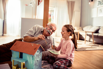 Grandfather and granddaughter painting a toy house together at home - Powered by Adobe