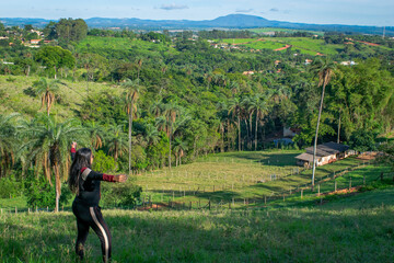 Woman contemplating the view from the top of a hill, a small farm in the Igarapé region, with lots of vegetation and mountains around. Minas Gerais Brazil.