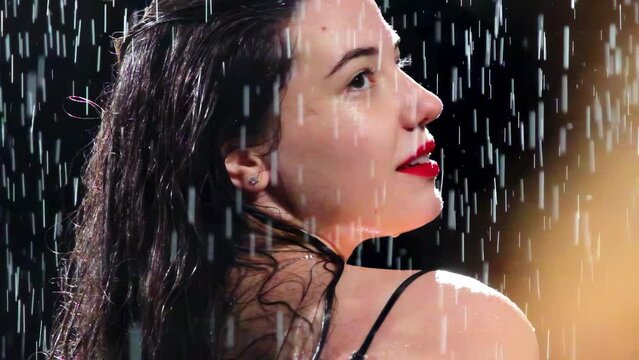 Pretty young woman smiles modestly and looks away sitting in rain. Attractive lady in black dress with open back carefully considers and enjoys fresh breath of wind