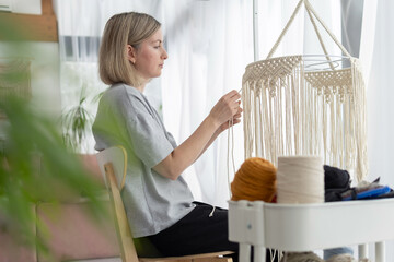 Middle-aged woman knits lampshade for interior using macrame technique. Woman knits boho chandelier...