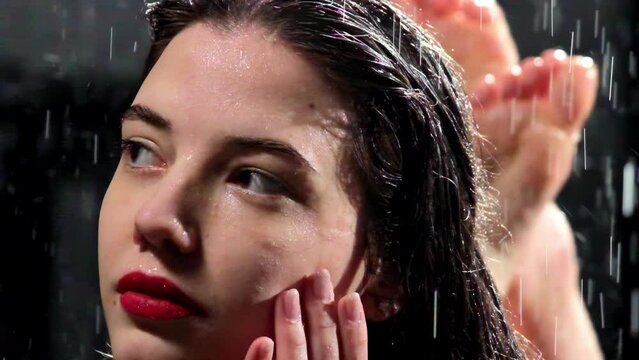 Lady lies with bent legs thoughtfully looking into distance in waterfall. Nice young woman with brunette wet hair thinks over problems in rain slow motion